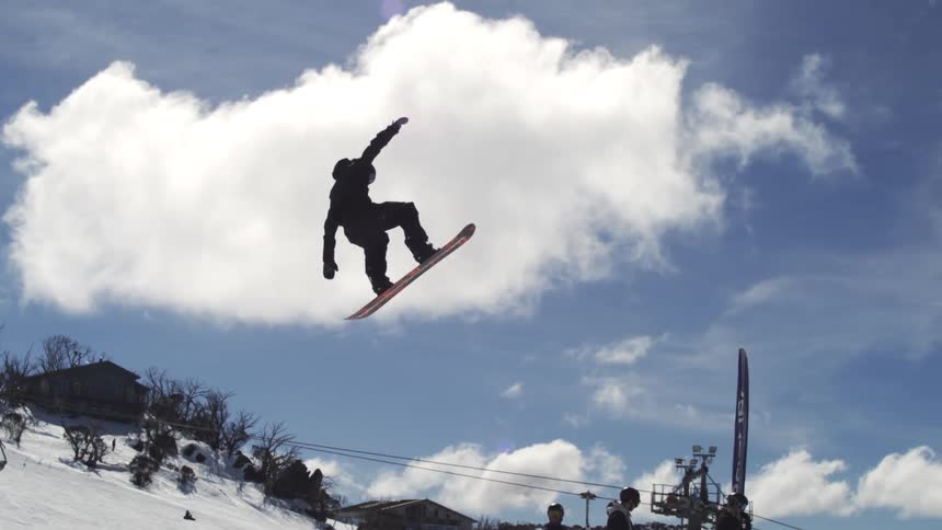 A snowboarder in dark winter clothing is performing a jump and spinning in the air with other people and ski lifts visible on a snowy slope below him