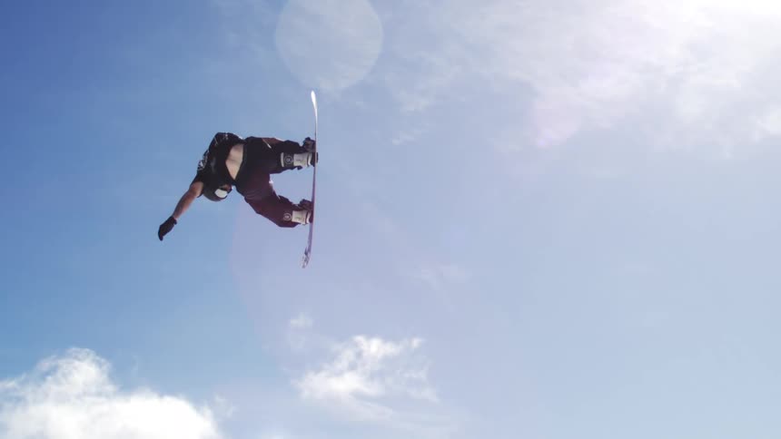 The video shows two shots of a snowboarder in midair on a snowy mountain the first shot is a closeup against a bright blue sky while the second pulls back to show the surrounding ski lifts and trees the words filmsupply are faintly visible in the