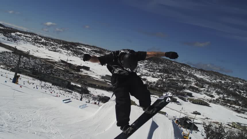 A snowboarder in black stands on a snow covered slope at a ski resort under a clear blue sky