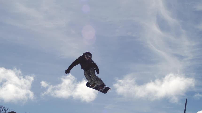 A snowboarder in dark winter clothing is performing a jump and spinning in the air against a bright blue sky with white clouds at a ski resort