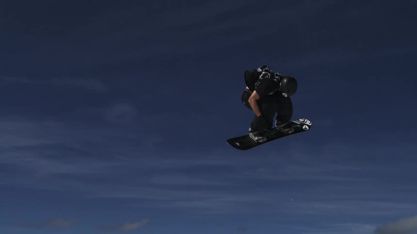 A male snowboarder in dark winter clothing is performing a jump and spinning in the air against a bright blue sky with the snowcovered slopes and a resort village visible in the background