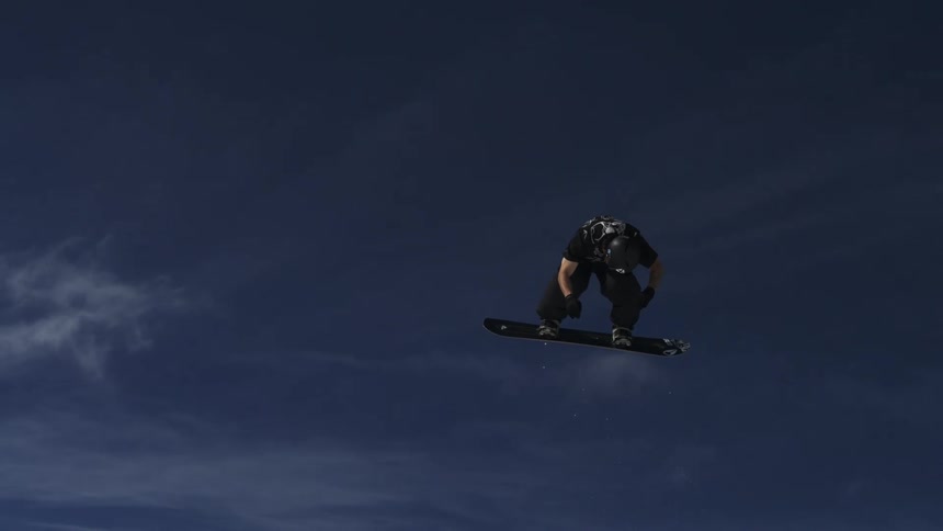 A snowboarder is performing an acrobatic trick off a jump and spinning in the air against a bright blue sky with a ski resort and mountains visible below him