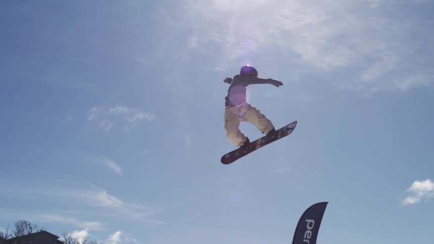 The video shows two shots of a snowboarder in midair on a snowy mountain the first shot is a closeup against a bright blue sky while the second pulls back to show the surrounding ski lifts and a perisher flag the words filmsupply are faintly vis