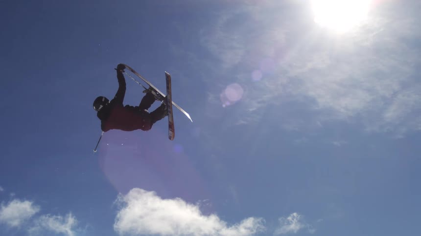 A skier performs an inverted aerial trick against a bright blue sky with scattered clouds with a ski resort visible in the background