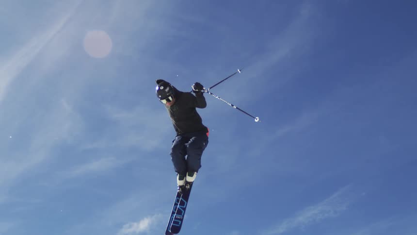 A male skier in dark winter clothing is performing a jump and spinning in the air with a snowcovered mountain and a bright blue sky in the background