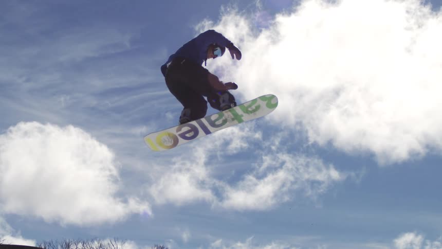 The video shows two shots of a snowboarder in midair on a snowy mountain the first shot is a closeup against a bright blue sky while the second pulls back to show the surrounding ski lifts and people the words filmsupply are faintly visible as a w