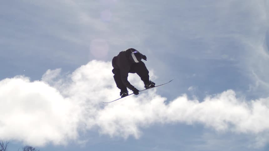 Snowboard rider in full snowboarding gear soaring high above the snowy mountain on a sunny day performing multiple somersaults in midair during an impressive stunt