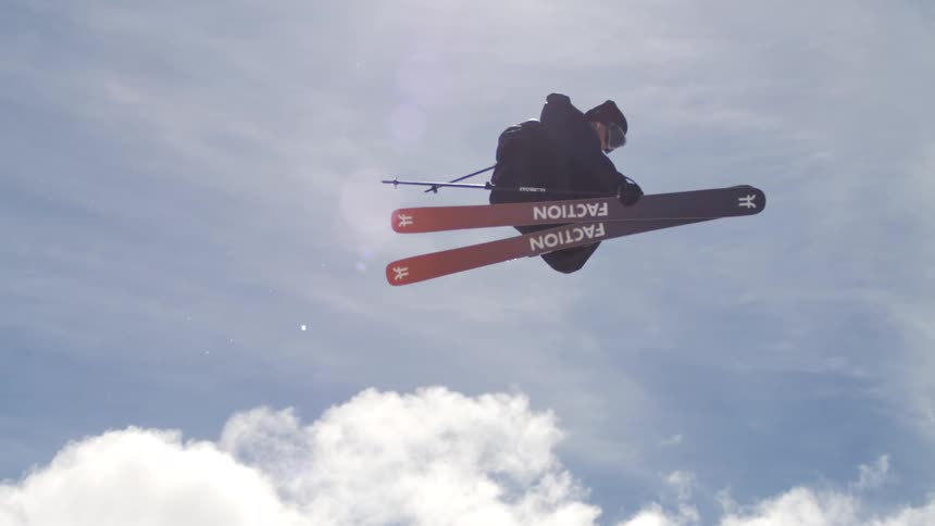 A male skier in dark winter clothing is performing a jump and spinning in the air with a snowcovered mountain and a bright blue sky in the background