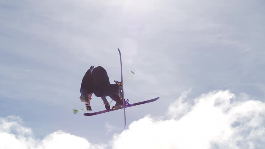 A skier in black performs an aerial trick with skis crossed and poles extended against a bright blue sky with clouds