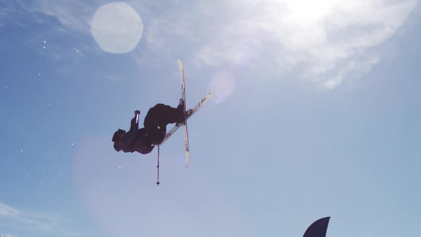 The video shows a skier performing a midair trick at a ski resort they are silhouetted against a bright blue sky as they flip and twist with their skis crossed in the background other skiers are visible on the slopes and there are buildings and a sk