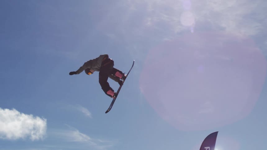 A male snowboarder is performing an acrobatic trick high in the air against a bright blue sky with the sun creating a lens flare and the snowy ground far below
