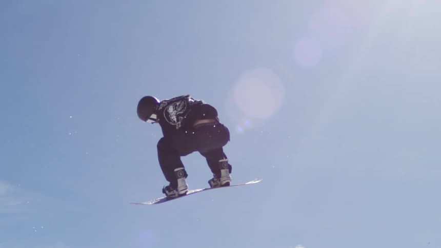 A snowboarder is performing an acrobatic trick high in the air with a snowcovered mountain and a bright blue sky with white clouds in the background