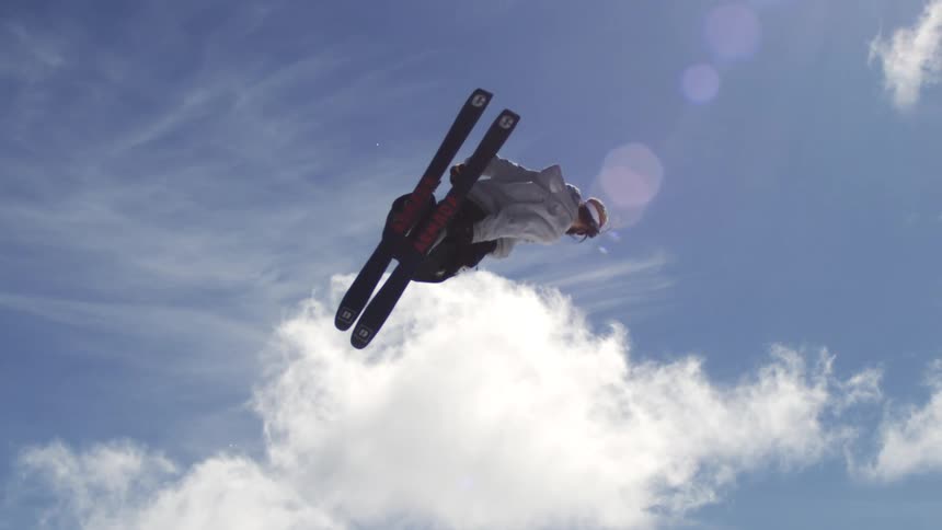 A skier is performing an acrobatic trick high in the air with the ski lift and other skiers visible in the background against a bright cloudy sky