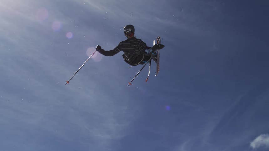 Man wearing a helmet and ski gear flung into the air while holding two ski poles midjump on a sunny winter day in the snowy mountains