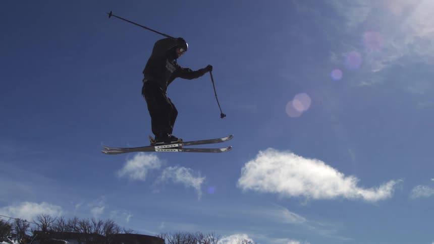 A male skier is performing an acrobatic trick high in the air with the ski lift and other skiers visible in the background against a bright cloudy sky