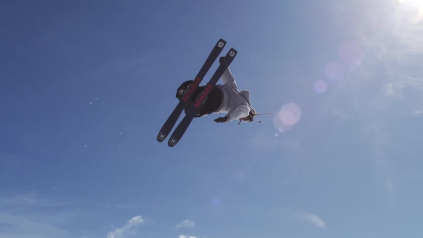 A skier wearing a black hoodie and white pants is performing a trick in the air with a bright sky in the background with the ski lift and other skiers visible in the distance