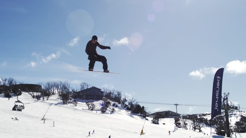 A snowboarder is performing an acrobatic trick off a jump in the air against a bright blue sky with a ski resort and mountains visible below him
