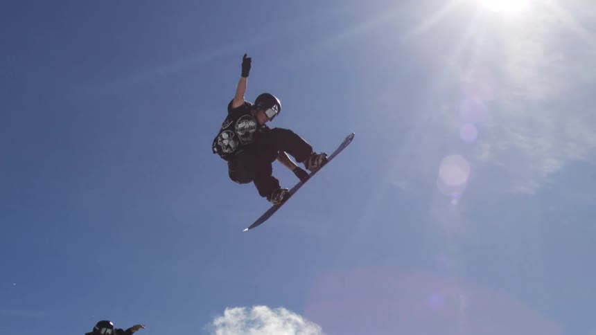 A snowboarder performs an aerial trick against a bright blue sky with scattered clouds showcasing a ski resort in the background