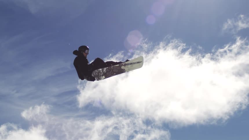 Snowboard rider in full snowboarding gear soaring high above the snowy mountain on a sunny day executing an impressive midair trick with the board angled dynamically