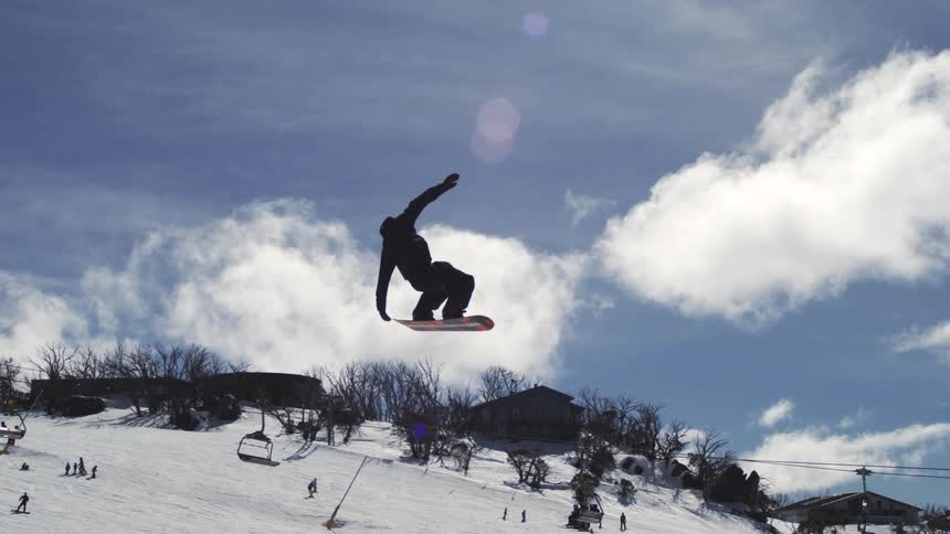 A male snowboarder in dark winter clothing is performing an acrobatic trick off a jump and spinning in the air against a bright blue sky with a ski resort and mountains visible in the background