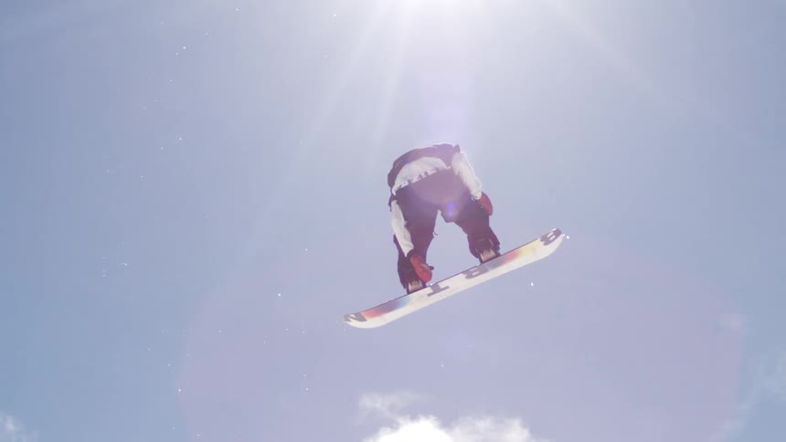 A snowboarder in black pants and a white jacket performs a trick in the air the shot is from a low angle showing the bright blue sky and some clouds behind the snowboarder with the snowy mountain and ski lifts visible below