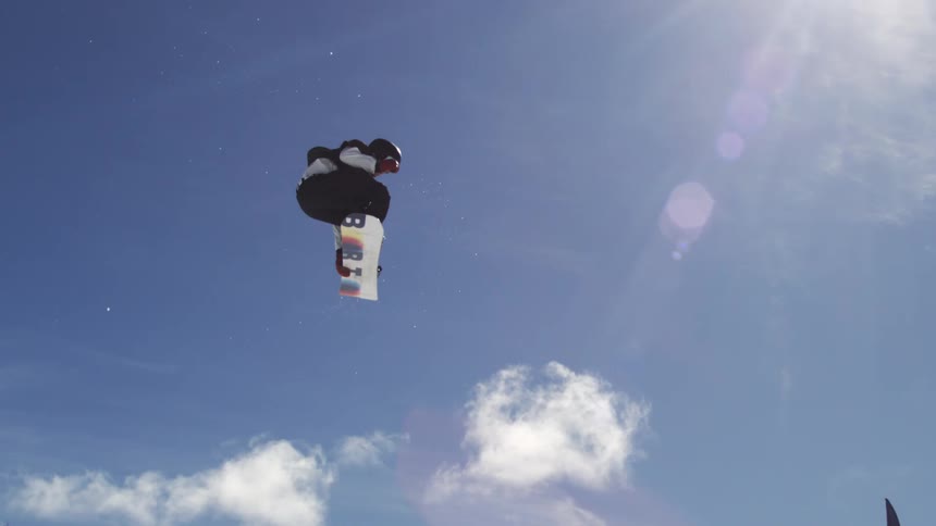 The video shows two shots of a snowboarder in midair on a snowy mountain the first shot is a closeup against a bright blue sky while the second pulls back to show the surrounding ski lifts and other people the words filmsupply are faintly visible