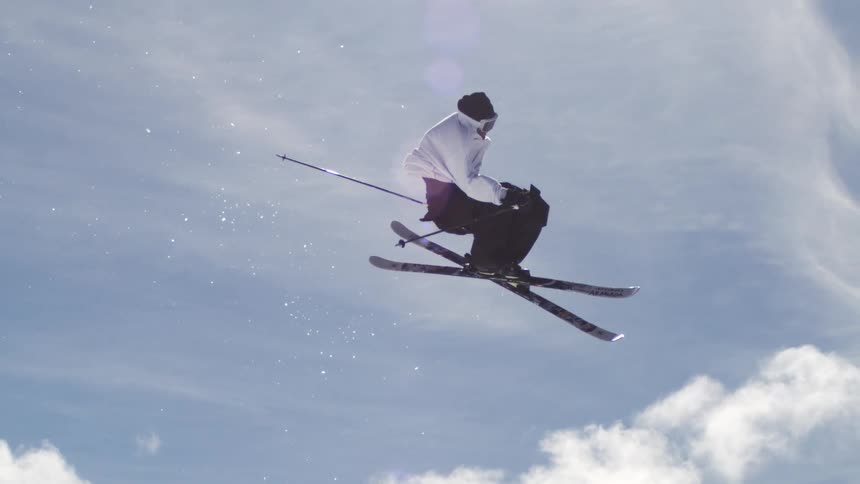 A male skier is performing an acrobatic trick high in the air with the ski lift and other skiers visible in the background against a bright cloudy sky