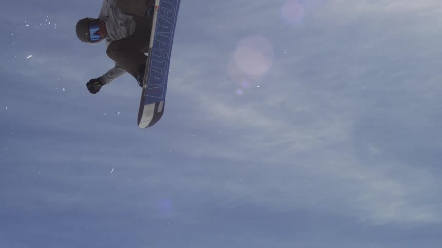 Snowboard rider in full snowboarding gear launching into the air at midday under a bright sunny sky performing a dynamic stunt high above the snowy mountain slopes