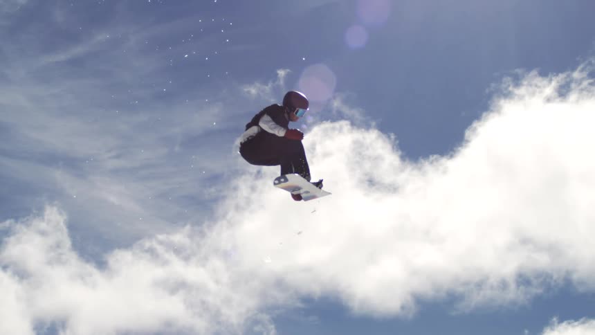 Snowboard rider in full snowboarding gear launching high into the air at midday on a sunny winter day in the mountains performing a double flip followed by a spin before landing on the snowy slope
