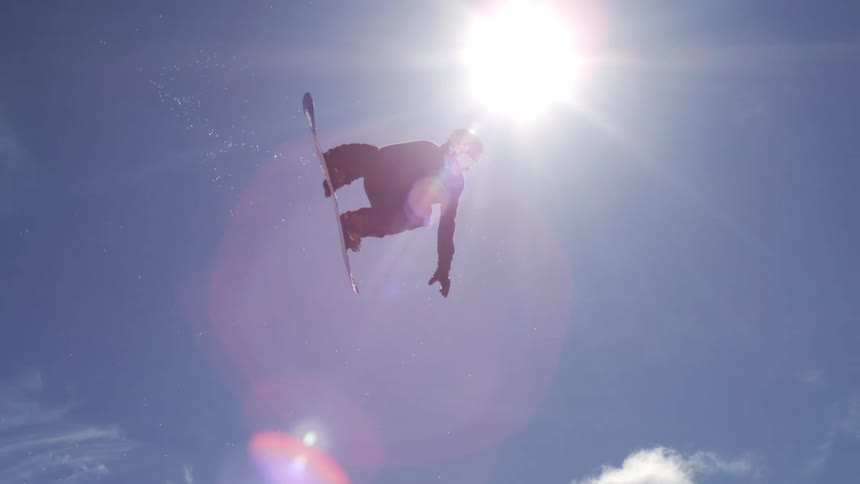 A male snowboarder in dark winter clothing is performing an acrobatic trick high in the air against a bright blue sky with the sun creating a lens flare and the snowy ground of a busy ski resort visible far below