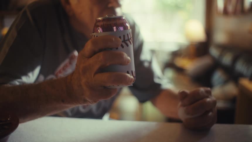 Elderly man in his living room at midday holding a beer can in one hand and a baseball in the other watching a game on tv with an excited animated expression