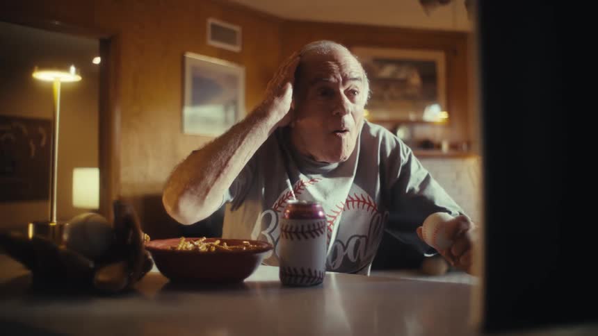 An elderly man wearing a baseball shirt and holding a baseball is watching a game on television reacting with a mix of excitement and surprise with a bowl of snacks on the table in front of him