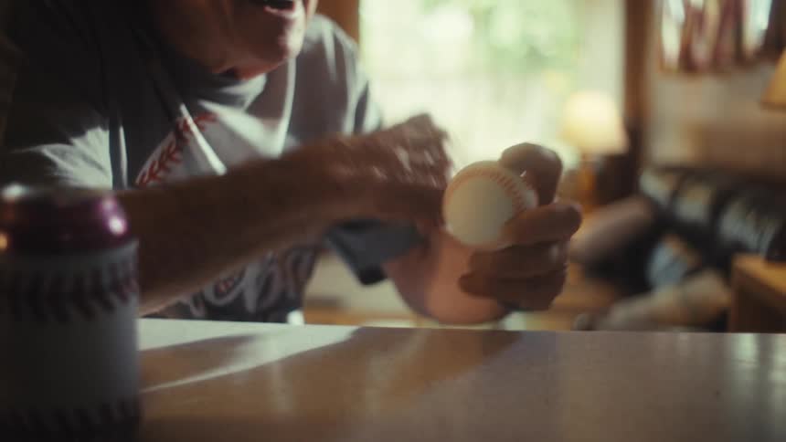 A man in a baseball shirt is holding a baseball at a table in a dimly lit room with his arm moving in a celebratory gesture