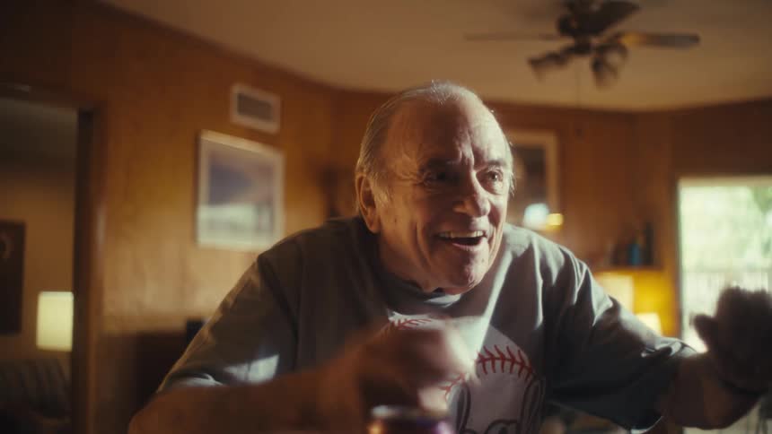 A shot of a joyful elderly man with a baseball in his hand cheering excitedly while watching a game offcamera he has a wide smile and appears to be in a welllit living room celebrating