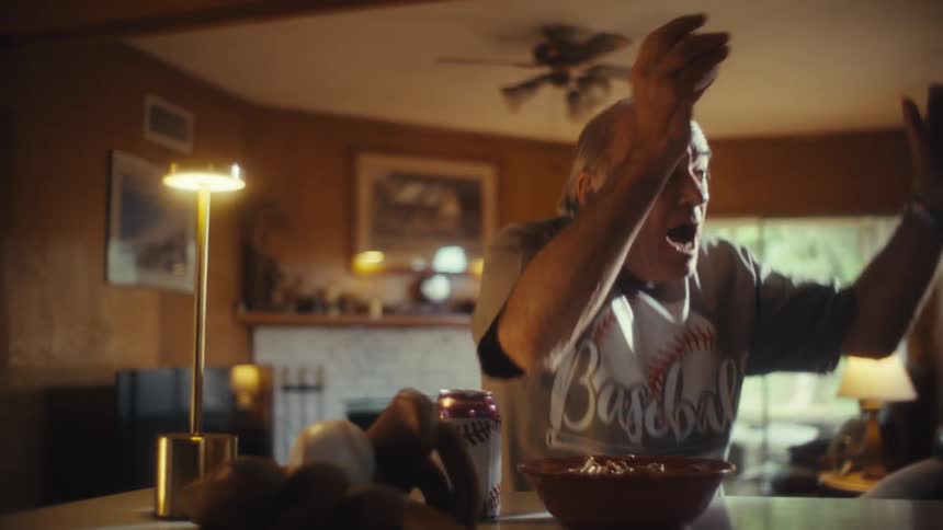 An elderly man wearing a baseball shirt and holding a baseball is watching a game on television reacting with a mix of excitement and disappointment with a bowl of snacks on the table in front of him