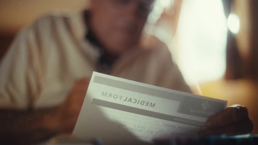 A shot of an older mans hands signing a medical form the text medical form is visible in reverse suggesting a reflection in a mirror he then picks up a brochure for fsp health and reads it carefully