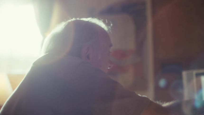 An older man is shown in a side profile shot with the sun streaming in from a window behind him casting his face in a bright light he raises his arm and gestures offscreen as if to a television or another person