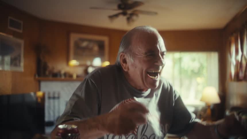 An elderly man in a baseball shirt watches a game intently from his kitchen counter he holds a baseball and suddenly cheers with a loud excited expression smiling as he pumps his fist