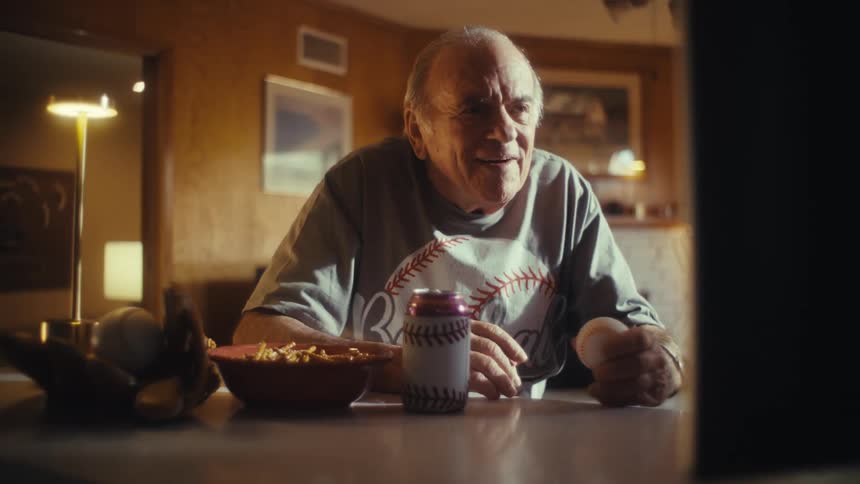 An elderly man in a baseball shirt celebrates a play while watching a game laughing and clapping as he holds a baseball and drinks a canned beverage