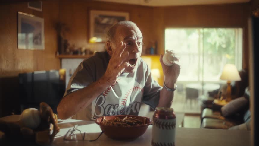 An elderly man sits at a kitchen counter holding a baseball in his hand as he watches a game on a nearby tv he looks on with joy and excitement a wide smile spreading across his face as he laughs and raises his fists in a moment of celebration