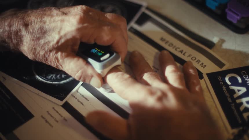 Elderly person removes a fingertip medical sensor lying on a counter covered in medical documents