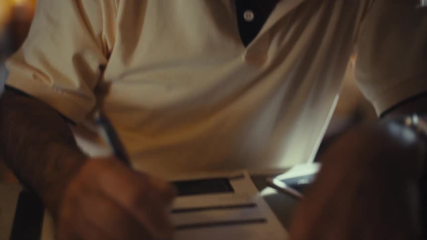 Elderly man indoors at midday wearing glasses and a lightcolored tshirt writing on a document with a pen