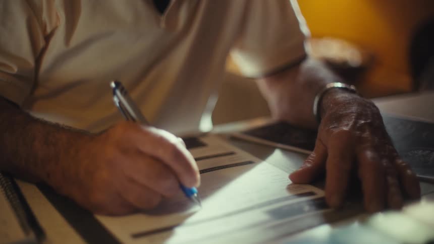 Elderly man indoors at midday wearing glasses and a lightcolored tshirt writing on a document with a pen