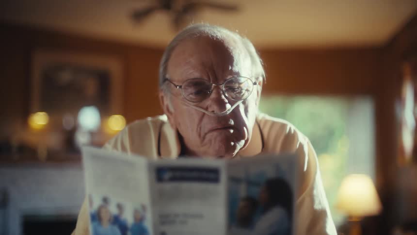 A man with a nasal cannula is shown in a closeup shot as he holds and reads a medical brochure he stares intently at the page before looking up at someone offscreen