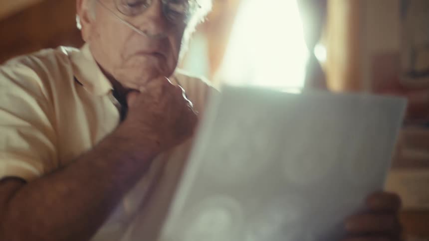 An elderly man with glasses and a nasal cannula holds up a brain scan and looks at it intently