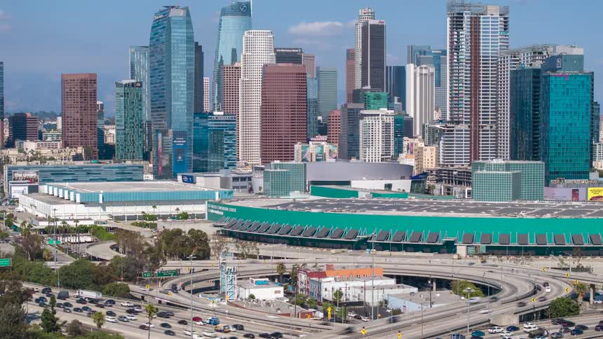 Los Angeles skyline on a sunny day bordered by highrise buildings and convention center with busy highway cuts through the scene packed with traffic flowing steadily beneath the bright blue sky