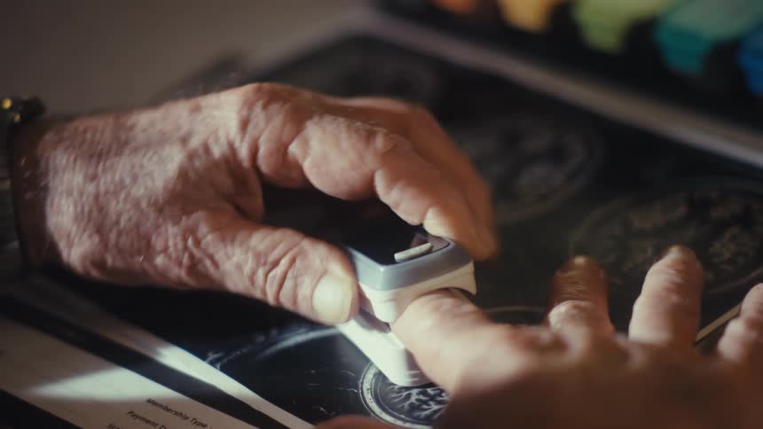 A close view shows a persons wrinkled hands with a pulse oximeter clipped to an index finger while taking a reading