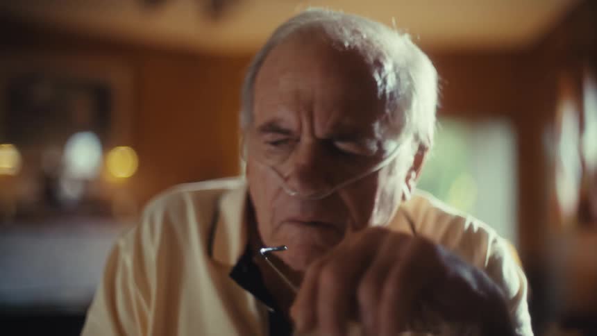 Elderly man wearing a nasal cannula seated indoors and writing with a pen on a document he pauses midsentence gaze drifting as sadness overtakes him lost in thought