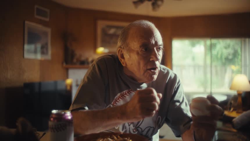 An elderly man in a baseball shirt sits at a kitchen counter holding a baseball in one hand he watches something offscreen then suddenly yells out with a joyful expression raising a fist in the air