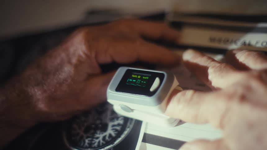 A close view of a persons fingers shows a pulse oximeter clipped to a finger and displaying a digital reading with the background of a medical folder out of focus
